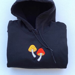 May include: Black hooded sweatshirt with a drawstring and embroidered mushroom design. The mushrooms are yellow and red with smiling faces. The sweatshirt is folded neatly on a white surface.