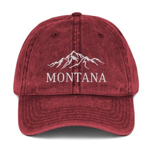 May include: A maroon baseball cap with a curved peak. The cap has white embroidered text reading "MONTANA" under a mountain range graphic. The cap has a vintage, worn appearance.