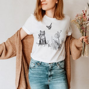 May include: White t-shirt featuring a black and white illustration of a cat sitting in grass with a butterfly and dandelions. The shirt has short sleeves and a crew neck.