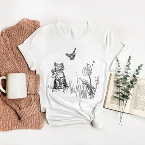 May include: White t-shirt featuring a black and white illustration of a cat, butterfly, and dandelions. The cat is wearing a scarf. A book, a mug, and a sweater are next to the shirt.