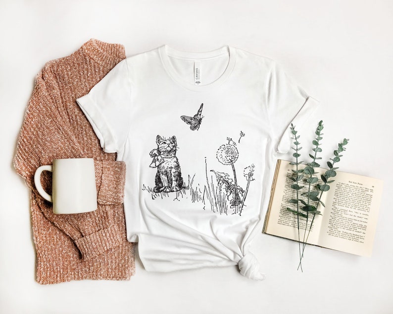 May include: White t-shirt featuring a black and white illustration of a cat, butterfly, and dandelions. The cat is wearing a scarf. A book, a mug, and a sweater are next to the shirt.