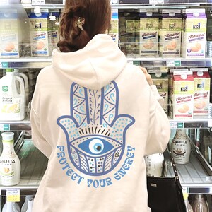 May include: A cream-colored hoodie with a blue Hamsa hand design and the text "PROTECT YOUR ENERGY." The design features an eye in the center of the hand. The hoodie is being worn in a grocery store.