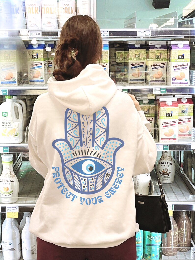 May include: A cream-colored hoodie with a blue Hamsa hand design and the text "PROTECT YOUR ENERGY." The design features an eye in the center of the hand. The hoodie is being worn in a grocery store.