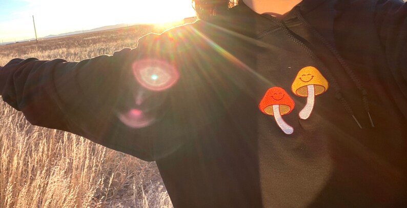 May include: Black hoodie with two embroidered mushroom patches. One mushroom is orange, the other is yellow, both with smiling faces. The background shows a field and the sun.