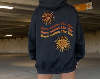 May include: Black hoodie with a retro sun graphic and the text "Here comes the Sun" in orange and yellow. The hoodie has a drawstring hood and a kangaroo pocket. The design is on the back of the hoodie.