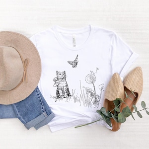 May include: White t-shirt with a black and white illustration of a cat, butterfly, and dandelions. A beige hat, blue jeans, and woven shoes are arranged around the shirt. The shirt has a crew neck and short sleeves.