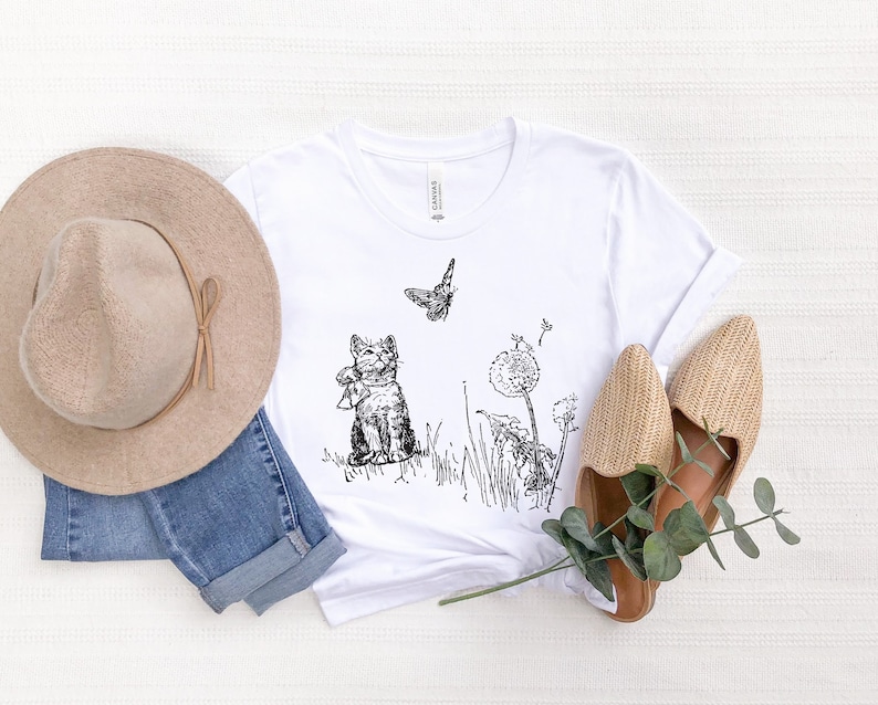 May include: White t-shirt with a black and white illustration of a cat, butterfly, and dandelions. A beige hat, blue jeans, and woven shoes are arranged around the shirt. The shirt has a crew neck and short sleeves.