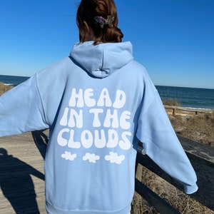 May include: A light blue hoodie with the text "HEAD IN THE CLOUDS" in white, along with three white cloud graphics. The hoodie is worn outdoors on a sunny day.