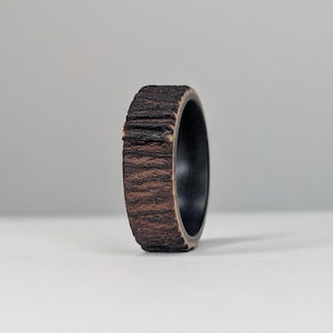 May include: A wooden ring with a dark brown, textured exterior and a black interior. The ring's exterior has a bark-like appearance, with visible ridges and a natural wood grain. The ring is standing upright against a neutral background.