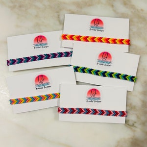 May include: Five woven friendship bracelets featuring chevron patterns in various colour combinations, each attached to a white display card with a circular logo that reads LJ Bracelet Boutique in black text with a pink and orange gradient circle.