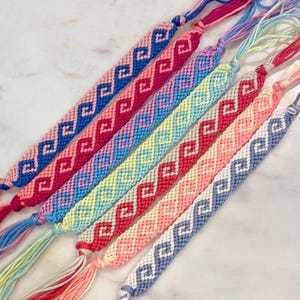 Puede incluir: Una colección de seis pulseras de la amistad tejidas a mano con un patrón geométrico de ondas repetitivo. Cada pulsera muestra una combinación única de dos colores, incluyendo hilo de bordar de algodón en azul, rosa, amarillo, rojo y blanco.