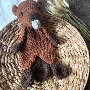 May include: A brown and tan crocheted beaver stuffed animal with a white nose. The beaver is sitting on a woven wicker basket.