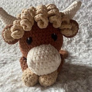 Peut inclure: Une peluche de vache des Highlands faite à la main au crochet. La peluche a un corps marron, des cornes crème et une crinière duveteuse brun clair. La vache a un museau blanc et des sabots brun clair. Les yeux sont noirs.