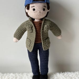 Peut inclure: Poupée crochetée représentant une personne portant une casquette de baseball bleue, une veste vert olive, une chemise marron et un pantalon bleu marine. La poupée a des chaussures noires et un teint clair.