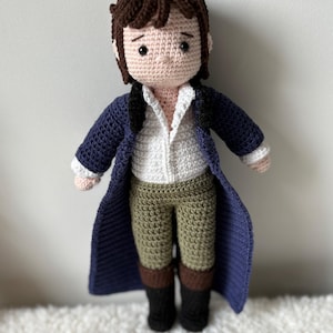 Könnte beinhalten: Handgefertigte Häkelpuppe, die eine Figur mit braunen Haaren darstellt, die ein weißes Hemd, eine olivgrüne Hose, einen marineblauen Mantel und schwarze Stiefel trägt. Die Puppe ist mit detaillierten Stichen gefertigt.