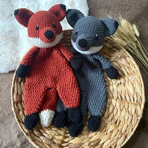 May include: Two crocheted stuffed animals, a fox and a wolf, are sitting in a woven basket. The fox is red with white accents and the wolf is gray with white accents.