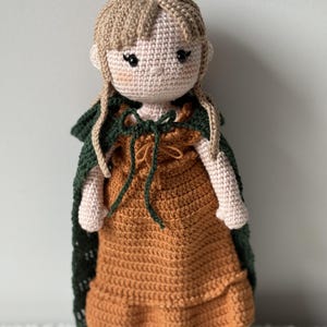 May include: A crocheted doll with long blonde hair, wearing a brown dress and a green cape. The doll has a sweet expression and is standing on a white surface.