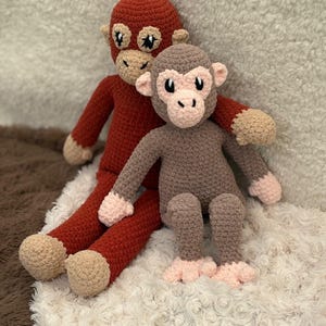 May include: Two handmade crochet monkey dolls. One is a deep red color, and the other is brown. Both have tan faces, ears, hands, and feet. The monkeys are sitting on a white, fluffy surface.