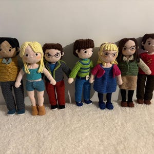 May include: A group of eight crocheted dolls, each with unique clothing and hairstyles. The dolls are standing on a white fluffy surface.