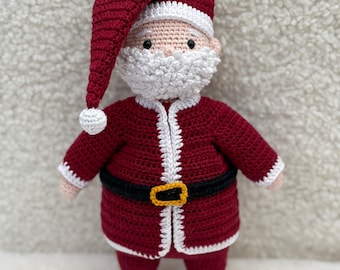 May include: A handmade crochet Santa Claus doll. The doll is primarily burgundy with white trim and a white beard. It has a black belt with a gold buckle and black boots. The Santa hat has a white pom-pom.