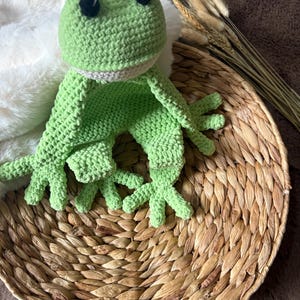 May include: A green crocheted frog toy with black eyes. The frog is sitting on a woven basket.