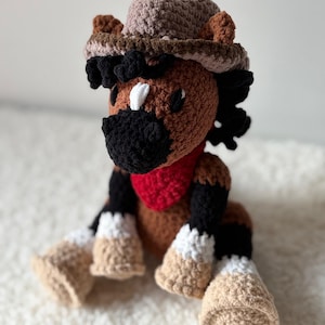 Peut inclure: Poupée cheval au crochet faite à la main, portant un chapeau de cowboy et un bandana rouge. Le cheval est marron avec une crinière et une queue noires, des marques blanches et des sabots beiges. Le chapeau est beige et marron.
