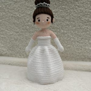 May include: A handmade crochet doll featuring a figure with light skin, rosy cheeks, and dark brown hair styled in a high bun with a grey tiara. The doll wears a strapless white gown and matching white elbow-length gloves, crafted with detailed yarn stitching.