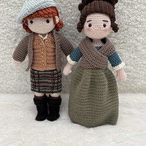 Peut inclure: Deux poupées crochetées à la main. L'une porte une veste marron, une jupe à carreaux et des bottes noires, avec un béret gris. L'autre poupée est vêtue d'une jupe verte, d'un haut bleu et blanc et d'un châle marron. Les deux poupées ont les cheveux bruns et se tiennent côte à côte.