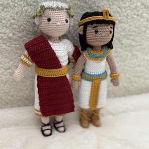 Peut inclure: Deux poupées crochetées à la main, l'une représentant un empereur romain et l'autre un pharaon égyptien. La poupée de l'empereur porte une toge rouge et blanche avec une couronne de laurier. La poupée du pharaon est dans une robe blanche avec des accents dorés et bleus.