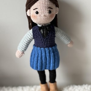 May include: A handmade crochet doll with brown hair and a light skin tone. The doll wears a blue skirt, a navy blue vest, a white shirt, black leggings, and tan boots. The doll has black eyes and a black bow tie.