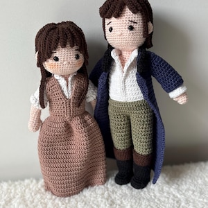 Puede incluir: Dos muñecos de ganchillo hechos a mano, una pareja, están uno al lado del otro. La muñeca de la mujer lleva un vestido rosa y la muñeca del hombre lleva un abrigo azul. Ambos muñecos tienen el pelo castaño y están hechos con una puntada texturizada.