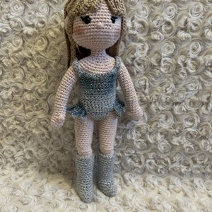 Puede incluir: Una muñeca de crochet con el pelo largo y rubio, que lleva un vestido azul brillante y botas plateadas. La muñeca está de pie sobre una superficie blanca y esponjosa.