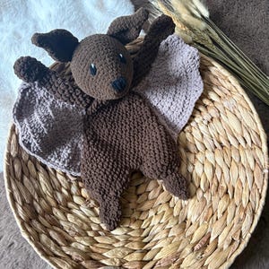 Puede incluir: Un juguete de murciélago de crochet marrón con un ala de crochet rosa. El juguete está sentado en una cesta tejida.