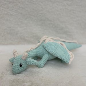 Peut inclure: Un dragon crocheté de couleur bleu sarcelle clair avec des accents crème. Le dragon a de petites cornes, des ailes et une longue queue. Les ailes et l'épine dorsale ont une bordure crème. Le dragon est posé sur une surface blanche et douce.