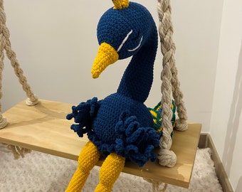 May include: A blue and yellow crocheted peacock plush toy sitting on a wooden swing. The peacock has a long neck and a yellow beak. The toy is sitting on a wooden swing with rope.