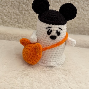 Peut inclure: Une figurine de fantôme crochetée en blanc portant une casquette noire avec des oreilles rondes. Le fantôme a des yeux et une petite bouche brodés en noir, et il porte un sac crocheté orange vif avec une bandoulière sur le corps.
