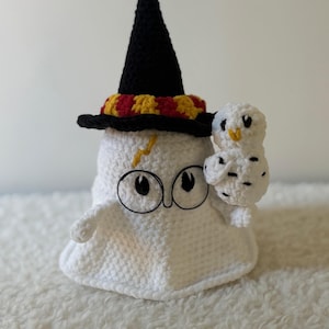 Peut inclure: Un fantôme blanc crocheté portant un chapeau de sorcier noir avec une bande rouge et or, et des lunettes rondes noires. Un petit hibou blanc est perché sur l'épaule du fantôme. Un éclair jaune est sur le front du fantôme.