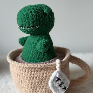 Peut inclure: Un dinosaure vert en crochet est assis dans une tasse à thé beige en crochet. La tasse a une anse et une étiquette de sachet de thé blanche avec "Tea" en noir. Le dinosaure a des dents blanches et de petits yeux noirs.