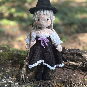 Peut inclure: Poupée crochetée à la main représentant une sorcière, avec un chapeau pointu noir, une robe noire à bordure blanche et un nœud violet. La poupée a des cheveux gris et tient un petit balai. La poupée est sur une bûche.