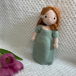 Pode incluir: Uma boneca de crochê com cabelo castanho comprido vestindo um vestido verde. A boneca está sentada em uma superfície branca texturizada com duas tulipas cor-de-rosa em primeiro plano.