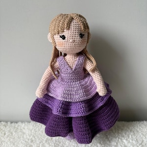 Peut inclure: Une poupée en crochet avec de longs cheveux bruns portant une robe violette à jupe à étages.