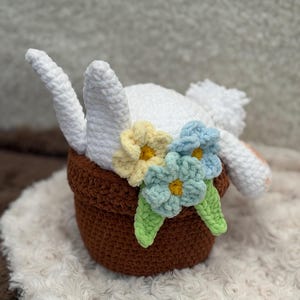 Peut inclure: Une tête et des oreilles de lapin en crochet blanc sortant d'un pot en crochet marron, ornés de fleurs en crochet jaunes, bleues et vertes. Le lapin repose sur une surface blanche et moelleuse, créant une esthétique fantaisiste et faite à la main.
