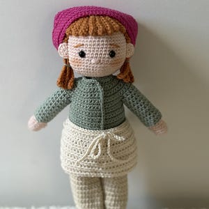 Peut inclure: Poupée au crochet faite à la main, avec des cheveux roux, portant un béret rose, une veste verte, un tablier crème et un pantalon beige. La poupée a des chaussures marron et se tient sur une surface blanche. Elle a des yeux noirs et une expression souriante.
