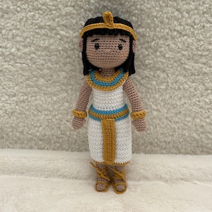 Peut inclure: Poupée crochetée représentant un pharaon égyptien. La poupée porte une robe blanche avec des détails dorés et bleus, une coiffe dorée et des cheveux noirs. La poupée mesure environ 20 cm de haut.