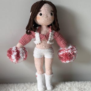 May include: A handmade crochet doll dressed as a cheerleader. The doll has brown hair, rosy cheeks, and is wearing a pink and white outfit with matching pom-poms. The doll is wearing white boots and shorts.