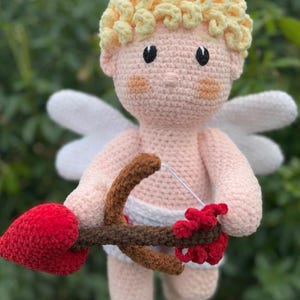 Peut inclure: Poupée Cupidon au crochet avec un corps pêche, des cheveux blonds bouclés et des ailes blanches. La poupée tient un arc marron et une flèche rouge en forme de cœur, idéale pour la Saint-Valentin.