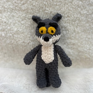 Peut inclure: Une poupée loup au crochet faite à la main dans les tons de gris, de blanc et de jaune. Le loup a une expression amicale avec de grands yeux jaunes, une poitrine blanche et un nez noir. La poupée est sur une surface blanche texturée.