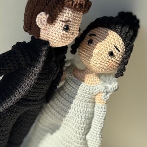 Puede incluir: Un conjunto de muñecas de crochet que representa a una pareja. El muñeco masculino lleva un traje negro y la muñeca femenina lleva un vestido blanco. Ambas muñecas tienen el pelo castaño y los ojos negros.