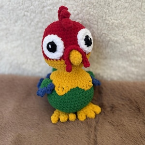 May include: A handmade crochet bird with a red head, yellow body, and green belly. The bird has large white eyes with black pupils, blue wings, and yellow feet. The plush toy is sitting on a brown surface.