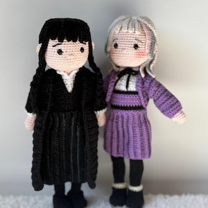 Peut inclure: Deux poupées crochetées à la main. L'une porte une robe et un manteau noirs avec des tresses noires. L'autre porte une robe et une veste violettes avec des cheveux gris. Les deux poupées ont des chaussures noires et se tiennent sur une surface blanche.